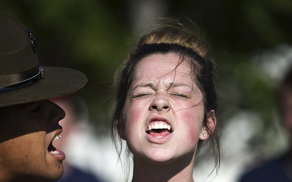A U.S. Marine Corps drill instructor in campaign cover and MARPAT camouflage uniform is shouting at a young female recruit wearing a Navy blue USMC-logoed t-shirt. The recruit stands at attention with eyes closed and mouth open, appearing to be responding or enduring the instruction. Additional recruits are visible but blurred in the background.