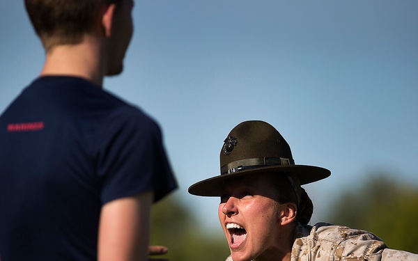 A U.S. Marine Corps drill instructor wearing the distinctive campaign cover (drill sergeant hat) and MARPAT camouflage uniform is shouting at recruits or civilians in navy blue t-shirts. The instructor is leaning forward aggressively in a classic drill instructor posture, mouth wide open mid-shout. The scene appears to take place outdoors during daylight hours, likely at a recruiting or training event.