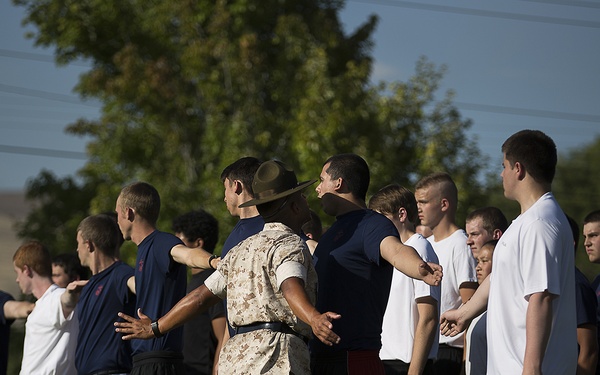 A military instructor or drill sergeant wearing desert camouflage uniform and a campaign cover (drill instructor hat) is organizing a group of young men in civilian athletic wear. The recruits or trainees are being arranged in a line formation outdoors on a grass field. The setting appears to be a military orientation, JROTC event, or recruiting activity.
