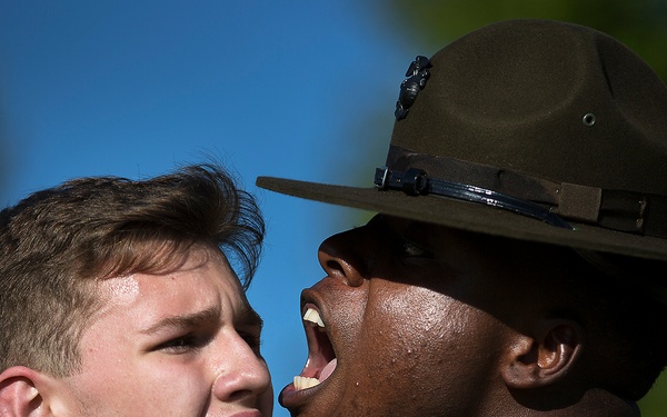 A military drill instructor wearing a campaign cover (drill sergeant hat) and desert MARPAT camouflage uniform is shouting directly into the face of a young recruit or civilian wearing a navy blue shirt. The recruit appears to be responding or reacting with a tense facial expression. This appears to be a classic military training or recruitment demonstration scenario.
