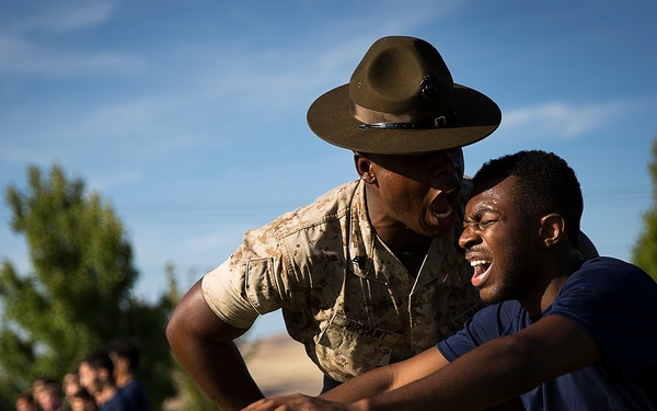 A U.S. Marine drill instructor in desert camouflage uniform and campaign cover (drill sergeant hat) is closely instructing or correcting a young recruit in civilian athletic wear during an outdoor training exercise. The recruit appears to be in a physical stance, possibly being coached on proper form or technique. A group of other recruits in dark athletic clothing stand in formation in the background.