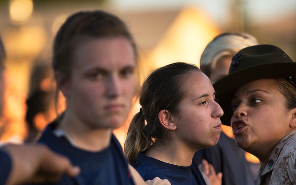 A female military drill instructor in Marine Corps camouflage uniform and campaign cover (drill sergeant hat) is giving instructions or corrections to a group of young female recruits wearing navy blue t-shirts. The recruits appear to be standing at attention with fists raised to their chests, suggesting a drill or physical training formation. The scene is lit by warm golden light consistent with early morning or late evening conditions.