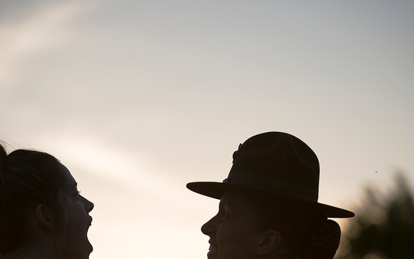 Two individuals are photographed in silhouette against a sunset sky, facing each other in close proximity. The backlit composition creates a dramatic contrast between the dark figures and the warm golden light of the setting sun. Trees are visible in the soft-focus background.