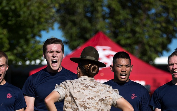 A female Marine drill instructor in desert camouflage uniform and campaign cover stands with hands on hips, facing four young male recruits wearing navy blue Marine Corps t-shirts. The recruits are shouting or responding loudly, appearing to be in a training or recruitment event setting. A red Marine Corps branded tent is visible in the background.