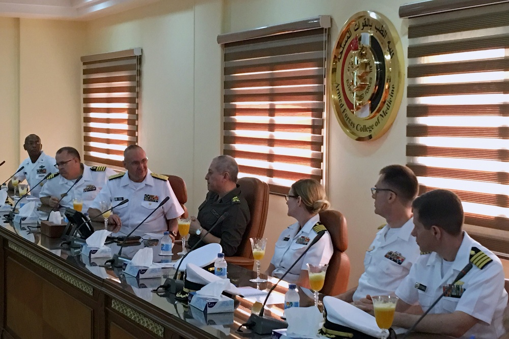 A formal military meeting is taking place at the Armed Forces College of Medicine, as indicated by the institutional seal visible on the wall. Several naval officers in white dress uniforms, along with at least one individual in olive/green military attire, are seated at a conference table. Orange juice drinks and water bottles are present on the table, suggesting a formal reception or briefing session.