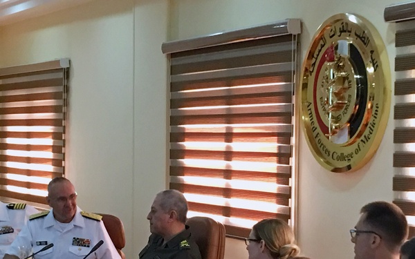 A formal military meeting is taking place at the Armed Forces College of Medicine, as indicated by the institutional seal visible on the wall. Several naval officers in white dress uniforms, along with at least one individual in olive/green military attire, are seated at a conference table. Orange juice drinks and water bottles are present on the table, suggesting a formal reception or briefing session.