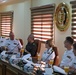 A formal military meeting is taking place at the Armed Forces College of Medicine, as indicated by the institutional seal visible on the wall. Several naval officers in white dress uniforms, along with at least one individual in olive/green military attire, are seated at a conference table. Orange juice drinks and water bottles are present on the table, suggesting a formal reception or briefing session.