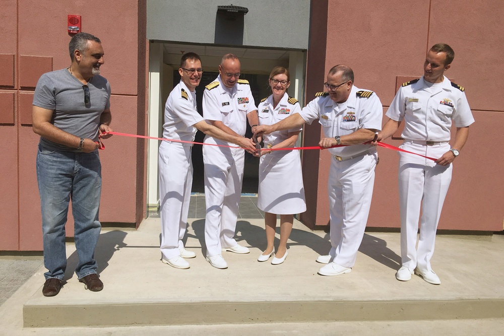A ribbon-cutting ceremony is taking place in front of a building, likely marking the opening or dedication of a military facility. Five individuals in white U.S. Navy dress uniforms and one civilian in casual clothing are participating in the ceremony. Multiple officers are jointly holding scissors to cut a red ribbon stretched across the entrance steps.