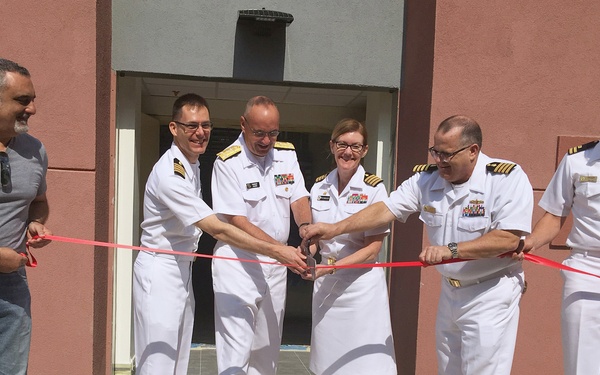 A ribbon-cutting ceremony is taking place in front of a building, likely marking the opening or dedication of a military facility. Five individuals in white U.S. Navy dress uniforms and one civilian in casual clothing are participating in the ceremony. Multiple officers are jointly holding scissors to cut a red ribbon stretched across the entrance steps.