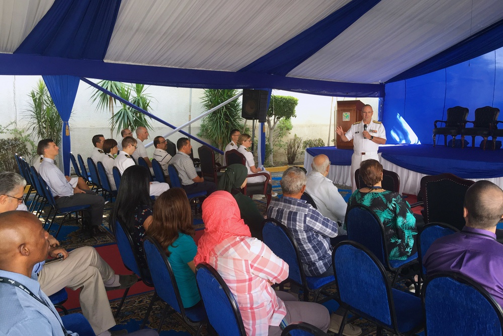 A formal outdoor event is taking place under a blue and white tent, where a military officer in white dress uniform is addressing an audience from a podium/stage area. The audience consists of a mixed group of civilians and military personnel seated in blue chairs. The setting appears to be an official ceremony or briefing, possibly at an embassy or military installation.