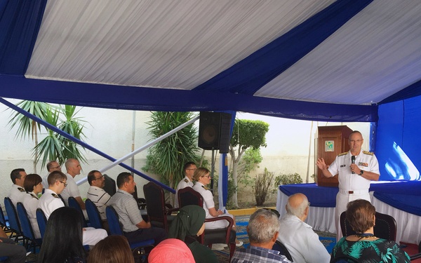 A formal outdoor event is taking place under a blue and white tent, where a military officer in white dress uniform is addressing an audience from a podium/stage area. The audience consists of a mixed group of civilians and military personnel seated in blue chairs. The setting appears to be an official ceremony or briefing, possibly at an embassy or military installation.
