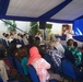 A formal outdoor event is taking place under a blue and white tent, where a military officer in white dress uniform is addressing an audience from a podium/stage area. The audience consists of a mixed group of civilians and military personnel seated in blue chairs. The setting appears to be an official ceremony or briefing, possibly at an embassy or military installation.