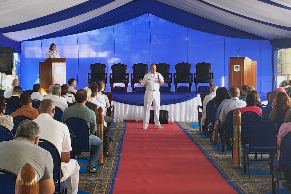 A formal military ceremony or briefing is taking place inside a large tent with blue and white decorations. A naval officer in white dress uniform is speaking to an audience while holding a microphone, with another uniformed individual at a podium on stage. The venue features a red carpet aisle, rows of chairs filled with attendees in both military and civilian attire.