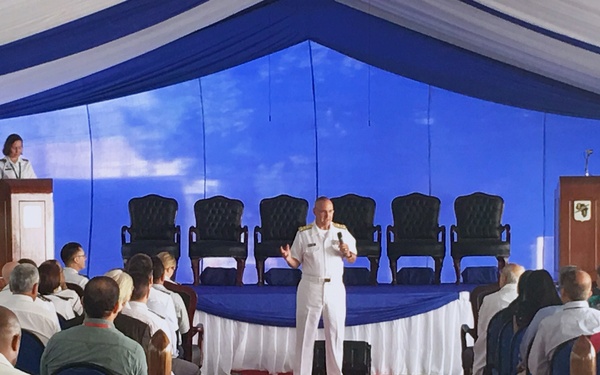 A formal military ceremony or briefing is taking place inside a large tent with blue and white decorations. A naval officer in white dress uniform is speaking to an audience while holding a microphone, with another uniformed individual at a podium on stage. The venue features a red carpet aisle, rows of chairs filled with attendees in both military and civilian attire.