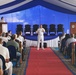 A formal military ceremony or briefing is taking place inside a large tent with blue and white decorations. A naval officer in white dress uniform is speaking to an audience while holding a microphone, with another uniformed individual at a podium on stage. The venue features a red carpet aisle, rows of chairs filled with attendees in both military and civilian attire.