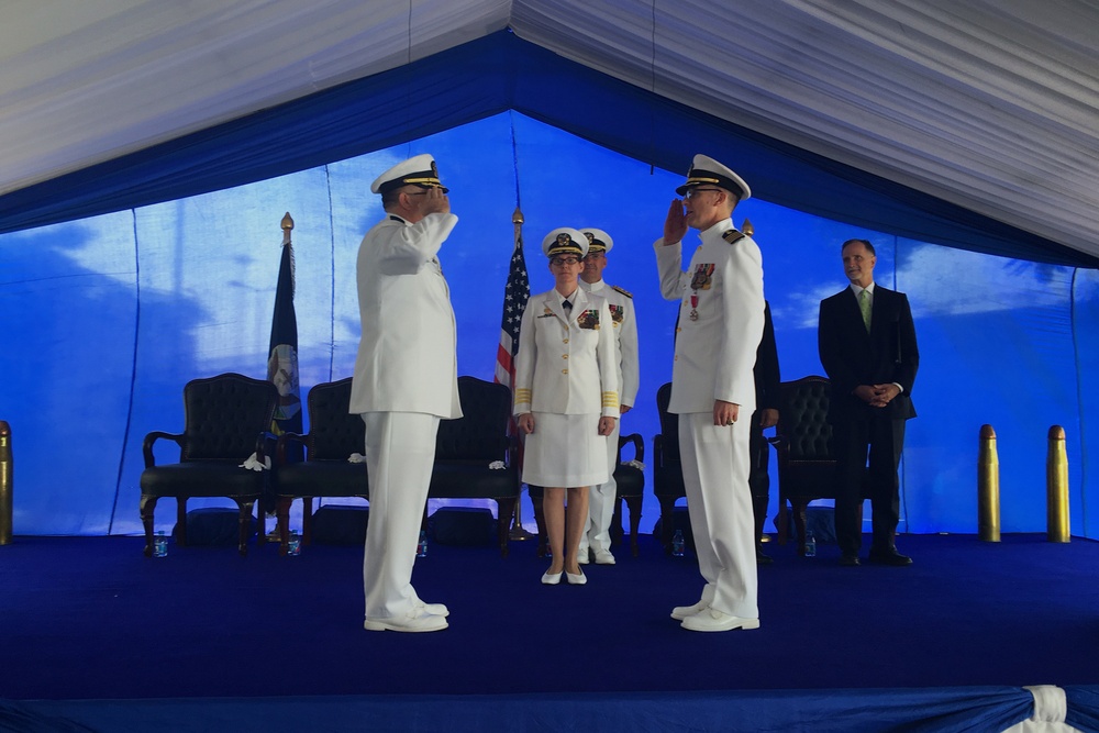 A formal military ceremony is taking place under a blue and white tent, featuring naval officers in white dress uniforms exchanging salutes. The scene appears to be a change of command or similar official military ceremony, with an American flag and what appears to be a unit flag visible in the background. Several officers and a civilian are present on a blue-carpeted stage.