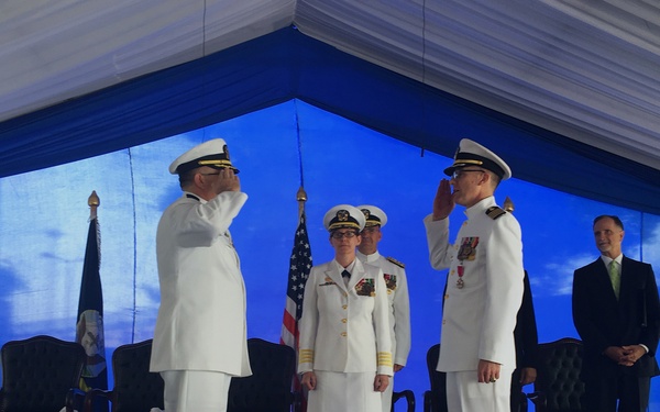 A formal military ceremony is taking place under a blue and white tent, featuring naval officers in white dress uniforms exchanging salutes. The scene appears to be a change of command or similar official military ceremony, with an American flag and what appears to be a unit flag visible in the background. Several officers and a civilian are present on a blue-carpeted stage.