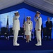 A formal military ceremony is taking place under a blue and white tent, featuring naval officers in white dress uniforms exchanging salutes. The scene appears to be a change of command or similar official military ceremony, with an American flag and what appears to be a unit flag visible in the background. Several officers and a civilian are present on a blue-carpeted stage.