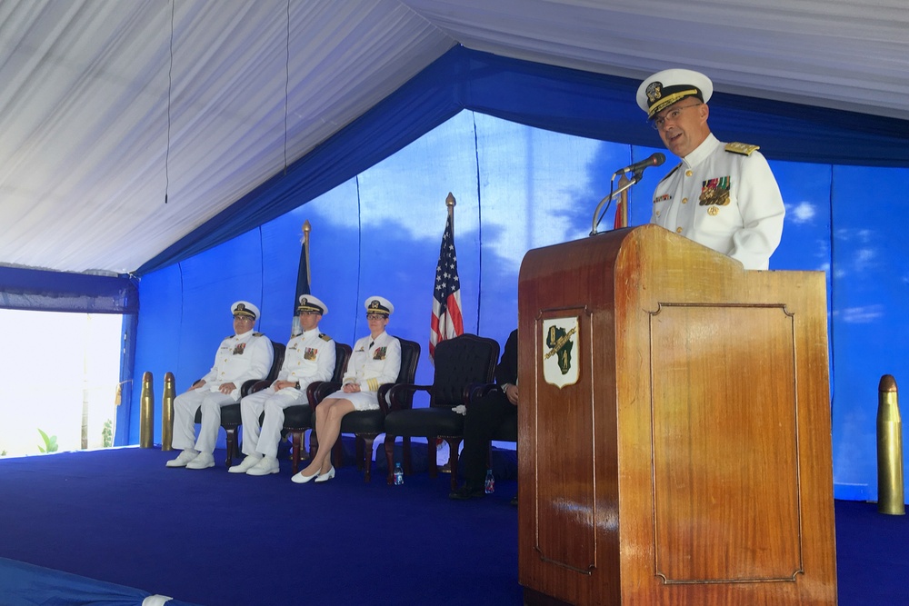 A formal U.S. Navy military ceremony is taking place under a blue and white tent. A senior naval officer in white dress uniform is speaking at a wooden podium, while three other naval officers in white uniforms are seated on a raised blue-carpeted stage behind him. An American flag and what appears to be a unit or command flag are displayed on the stage.