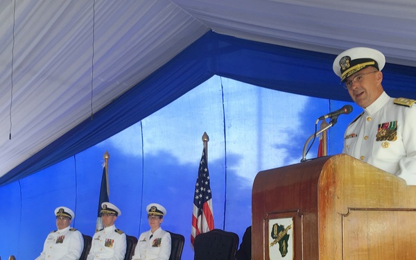 A formal U.S. Navy military ceremony is taking place under a blue and white tent. A senior naval officer in white dress uniform is speaking at a wooden podium, while three other naval officers in white uniforms are seated on a raised blue-carpeted stage behind him. An American flag and what appears to be a unit or command flag are displayed on the stage.