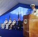 A formal U.S. Navy military ceremony is taking place under a blue and white tent. A senior naval officer in white dress uniform is speaking at a wooden podium, while three other naval officers in white uniforms are seated on a raised blue-carpeted stage behind him. An American flag and what appears to be a unit or command flag are displayed on the stage.