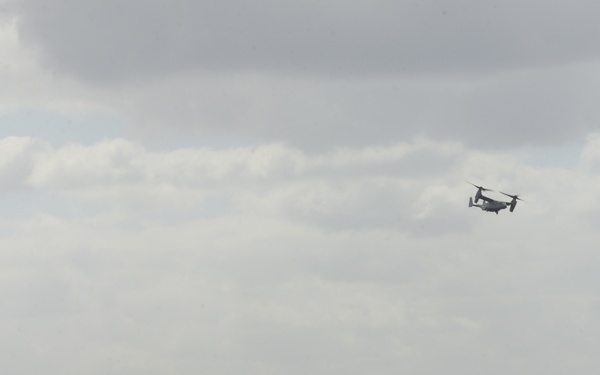 The image shows a large naval aircraft carrier at sea with an MV-22 Osprey tiltrotor aircraft flying nearby. The scene appears to be an open ocean environment under partly cloudy skies. The aircraft is positioned to the right of the carrier, suggesting an approach, departure, or patrol operation.