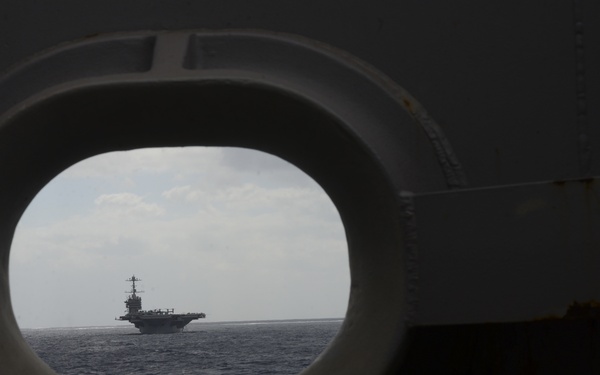 A large naval vessel, consistent with an aircraft carrier, is viewed through a circular porthole or hawse pipe opening from another ship. The carrier is visible at a distance on open ocean under a partly cloudy sky. The framing device creates a dramatic compositional contrast between the dark foreground structure and the bright seascape beyond.