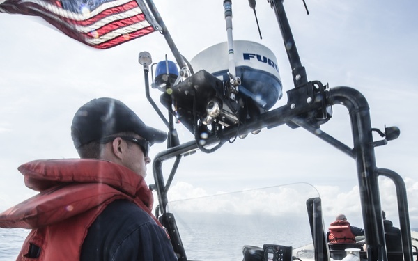 A person wearing a red life vest and black cap is operating a patrol or rescue boat on open water. The vessel is equipped with a Furuno radar dome, multiple control panels, and displays an American flag at the stern. A second person in a red life vest is visible in the background.