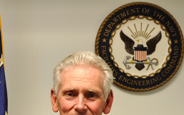 An older man with white hair is posing for what appears to be an official portrait or headshot. He is standing in front of a U.S. Navy Facilities Engineering Command seal mounted on a wall, with an American flag visible to his left. He is wearing a casual striped polo shirt and smiling at the camera.