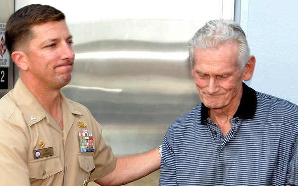 A U.S. Navy officer in tan service uniform is shaking hands with an elderly civilian man in front of an elevator. The naval officer is wearing multiple service ribbons and a name tag, suggesting a formal or ceremonial meeting. The interaction appears cordial, with the officer also placing a hand on the civilian's shoulder.