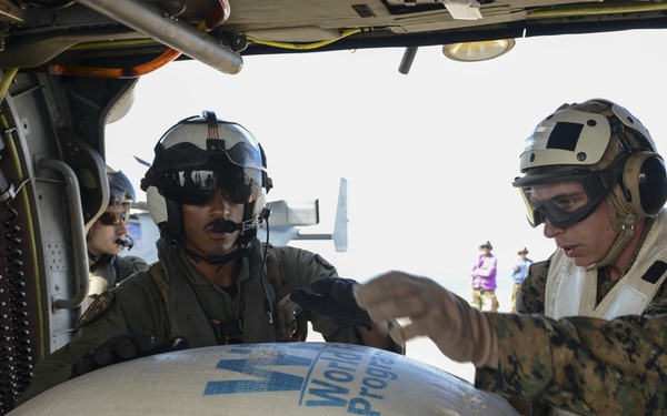 Military personnel, including what appear to be U.S. Marines and a helicopter crew member, are handling large sacks marked 'World Food Programme' and 'Rice' inside or alongside a military helicopter. The operation appears to be a humanitarian aid delivery mission, with WFP food supplies being loaded or unloaded. Civilians are visible in the background, suggesting an active aid distribution site.