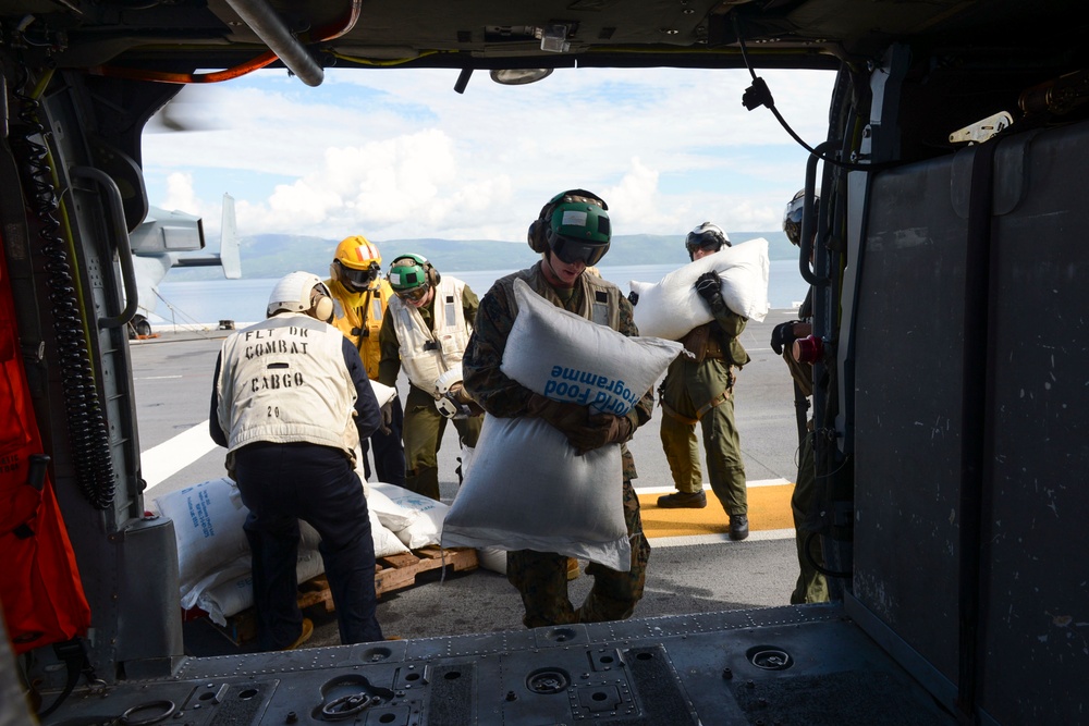 Military personnel are loading World Food Programme (WFP) supply bags onto a helicopter, likely as part of a humanitarian aid or disaster relief operation. The scene takes place on what appears to be a naval vessel flight deck, with personnel wearing flight deck gear and combat cargo vests. The operation involves transferring white sacks marked 'World Food Programme' into the open cargo bay of a helicopter.