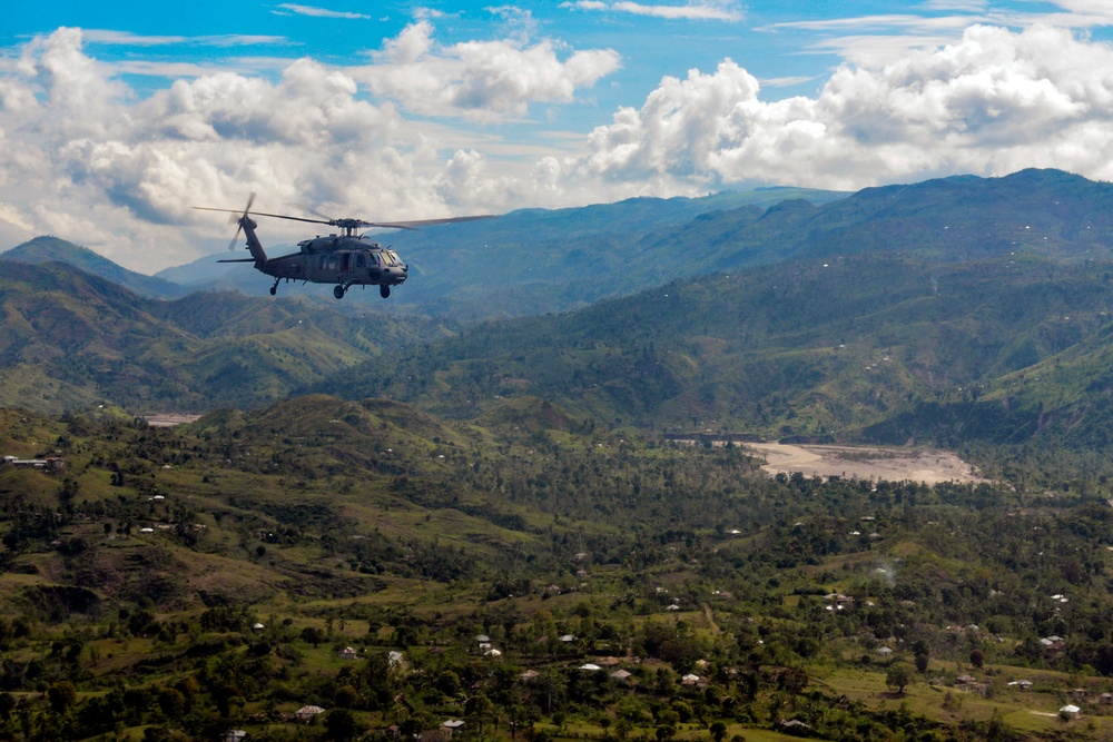 A military helicopter, consistent with a UH-60 Black Hawk, flies over a lush, mountainous tropical landscape with scattered rural settlements visible below. The terrain features rolling green hills, a river valley with a sandy riverbed, and sparse structures suggesting a developing or rural region. The sky is partly cloudy with bright blue patches above the mountain ridgeline.