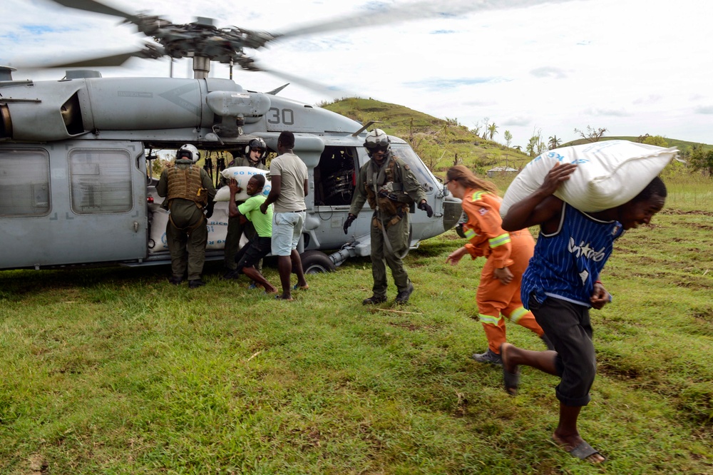 A military helicopter, marked with the number '30', has landed on a grassy field as part of what appears to be a humanitarian aid operation. Military personnel in flight suits and tactical gear are unloading large white sacks, likely food supplies marked '50 KG', with assistance from local civilians. A relief worker in an orange jumpsuit is also present, suggesting a coordinated disaster relief effort.