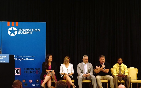 A panel discussion is taking place at a 'Transition Summit' event organized by 'Hiring Our Heroes' and the USO. Five panelists are seated on a stage while a moderator speaks at a podium on the left. The audience consists largely of military personnel in camouflage uniforms.