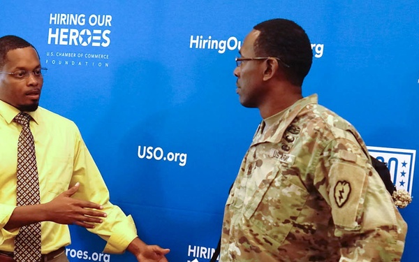 Two men are engaged in conversation in front of a 'Hiring Our Heroes' U.S. Chamber of Commerce Foundation step-and-repeat backdrop, which also displays USO.org branding. One man is dressed in business casual attire and is holding a paper sign reading 'Reserved for: Melvin Corey,' while the other is wearing U.S. Army camouflage (OCP/MultiCam) uniform with a 25th Infantry Division patch visible. The setting appears to be a formal event focused on veteran employment.