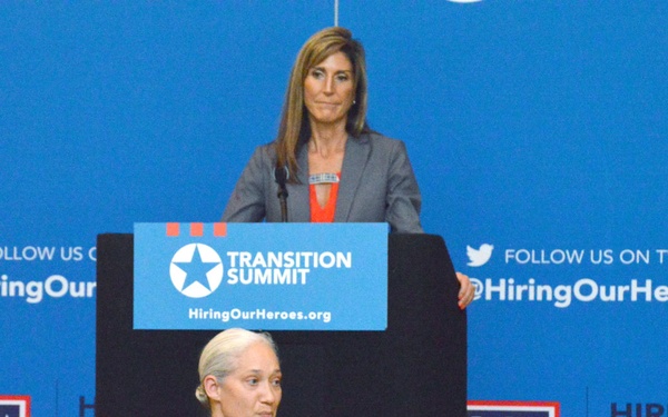 A military service member in camouflage uniform speaks into a microphone at a 'Transition Summit' event hosted by Hiring Our Heroes, a U.S. Chamber of Commerce Foundation initiative. A civilian woman in a gray blazer stands at the podium behind the speaker. The event appears to be focused on military-to-civilian employment transition, with USO branding also visible.