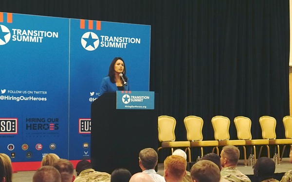 A woman in a blue blazer is speaking at a podium during a 'Transition Summit' event co-hosted by the USO and Hiring Our Heroes organizations. The audience consists predominantly of military personnel in camouflage uniforms seated in rows facing the stage. The event appears to be focused on helping service members transition to civilian employment.