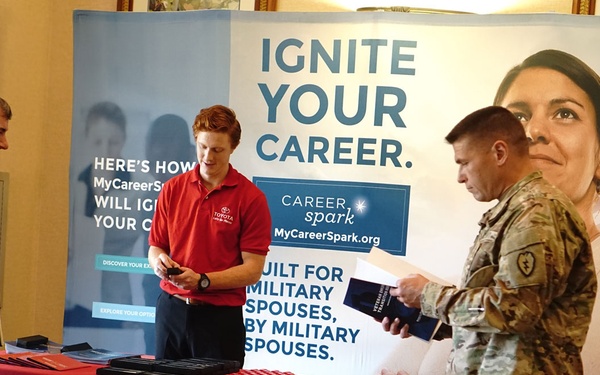 A Toyota-sponsored career fair booth targeting military spouses, featuring the 'Career Spark' program (MyCareerSpark.org). Two military personnel in camouflage uniforms are interacting with a civilian representative in a red Toyota polo shirt. The booth displays banners with the message 'Ignite Your Career' and 'Built for Military Spouses, by Military Spouses.'