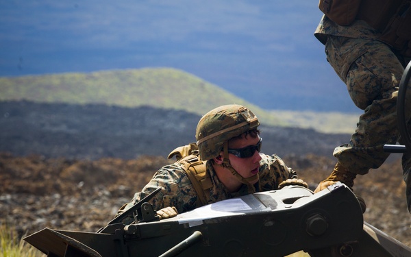 A U.S. Marine in full combat uniform and gear is working with heavy equipment, appearing to handle or inspect a bulldozer blade or similar attachment on rocky, volcanic terrain. A second Marine is partially visible operating or standing on the equipment. The setting appears to be a training exercise in a volcanic landscape, likely in Hawaii based on the distinctive dark lava rock terrain.