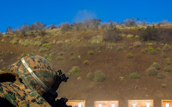 A military service member in digital camouflage (MARPAT pattern) is firing a rifle from a prone position on a shooting range. The shooter is aiming at a row of paper targets mounted on wooden frames downrange. Dust or smoke is visible near the muzzle, indicating active firing.