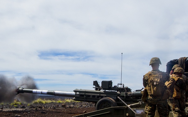 Military personnel are operating a self-propelled howitzer artillery system during a live-fire exercise on what appears to be a volcanic terrain. The weapon has just been discharged, as evidenced by the visible muzzle blast and smoke emanating from the barrel. Two or three soldiers in combat uniforms and tactical gear are positioned beside the artillery piece.