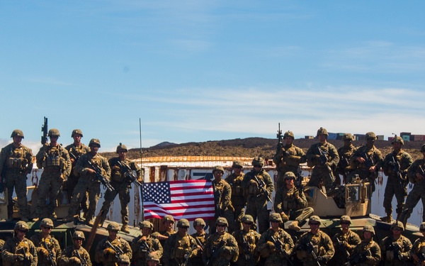 A large group of military personnel in camouflage uniforms pose for a unit photograph in an arid, rocky terrain. They are positioned around two military light armored vehicles, with several soldiers standing on top of the vehicles. A United States flag is prominently displayed at the center of the group.