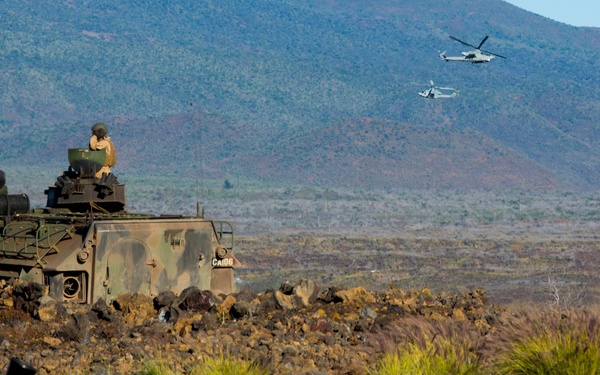 A military exercise is underway in a volcanic terrain landscape, likely in Hawaii based on the distinctive lava rock terrain. An armored personnel carrier with a soldier in the turret is positioned among lava rocks, while two military helicopters fly in the background over mountainous terrain. Equipment and gear are visible in the foreground near what appears to be a marked range or landing zone.