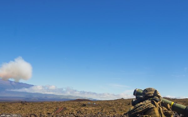 A military service member in camouflage gear is firing a shoulder-launched missile system from a prone/kneeling position on rocky volcanic terrain. The missile is visibly airborne in the upper left of the frame with a smoke trail behind it. The setting appears to be a training exercise on a volcanic landscape, likely in Hawaii based on the terrain and mountain visible in the background.