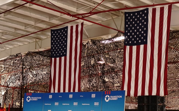 An indoor military event identified as a 'USO Transition Summit' and 'Hiring Our Heroes' program is taking place in what appears to be a large hangar or gymnasium. A speaker stands at a podium on a stage with a blue branded backdrop, addressing a large seated audience composed primarily of military personnel in uniform. Two large American flags and camouflage netting are displayed prominently as decorations.
