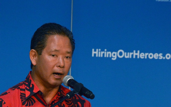 A man in a red and black patterned shirt is speaking at a podium during the 'Transition Summit' event organized by HiringOurHeroes.org. The event appears to be associated with the U.S. Chamber of Commerce Foundation, focused on helping military veterans transition to civilian employment. He is addressing an audience using a microphone mounted on the wooden lectern.