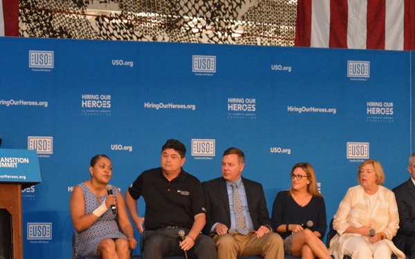 A panel discussion is taking place at a USO 'Transition Summit' co-hosted with 'Hiring Our Heroes' organization. A moderator stands at a podium on the left while six panelists are seated on stage in front of a branded blue backdrop. The audience, which includes military personnel in camouflage uniforms, is seated facing the stage.