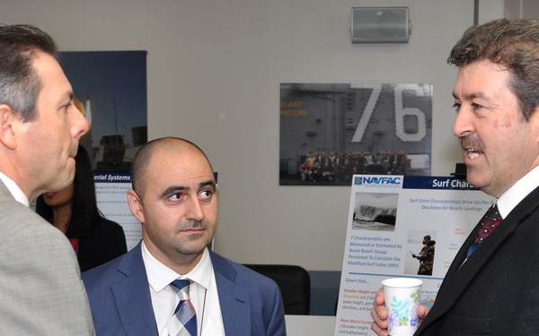 Three men in business attire are engaged in conversation at what appears to be a professional or military-related exhibition or briefing event. The man on the right is holding a disposable cup and appears to be speaking, while the other two listen. Display boards and posters related to NAVFAC (Naval Facilities Engineering Command) are visible in the background.