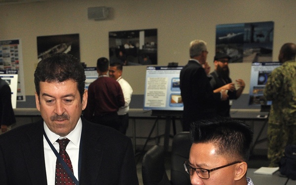 Two men in the foreground are engaged in a discussion while reviewing a display board or presentation material. The setting appears to be a professional conference, briefing, or technology exposition with multiple display boards and posters visible in the background. Several other attendees are visible in the background, including one individual in military camouflage uniform.